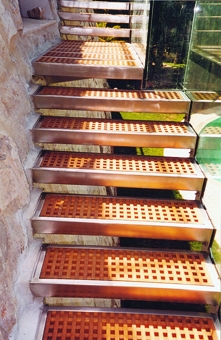 Scala in acciaio inox e pedate in legno pregiato grigliato. Il parapetto è in cristallo blindato.